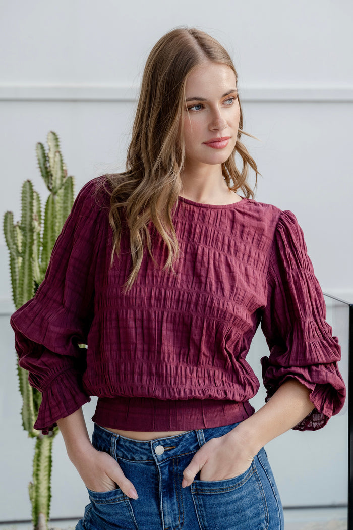 Woman wearing a burgundy long sleeve top with shirred detailing and fluted cuffs, styled with high-waisted blue jeans.