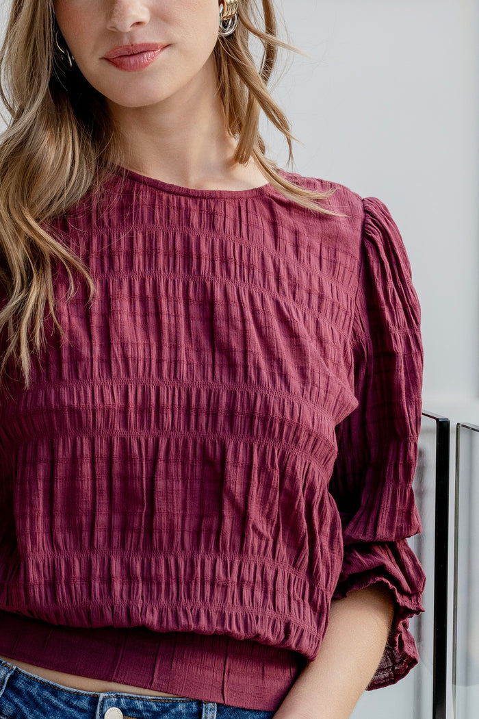 Woman wearing a burgundy long sleeve top with shirred detailing and fluted cuffs, styled with high-waisted blue jeans.