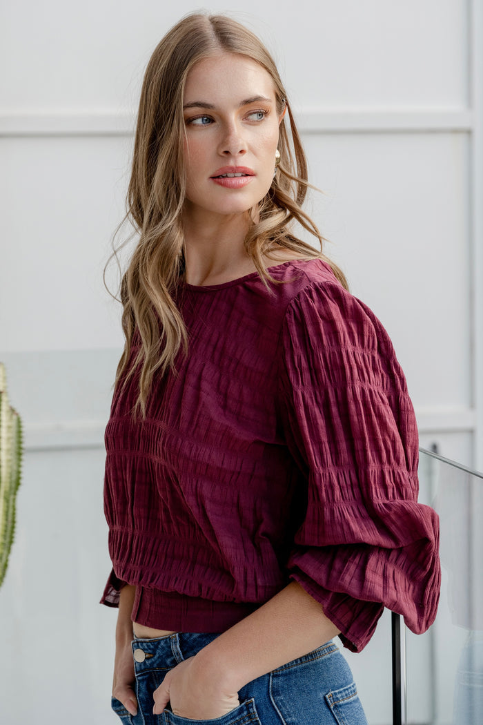 Woman wearing a burgundy long sleeve top with shirred detailing and fluted cuffs, styled with high-waisted blue jeans.