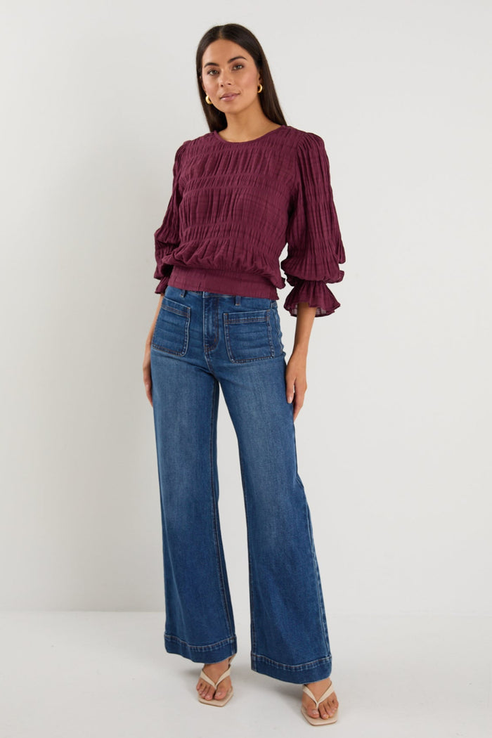 Woman wearing a long-sleeve textured blouse in deep plum, styled with blue high-waisted jeans and gold hoop earrings.