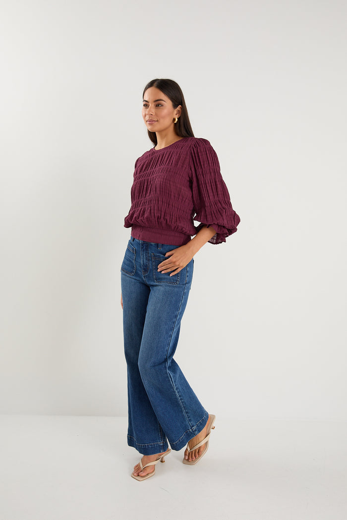 Woman wearing a long-sleeve textured blouse in deep plum, styled with blue high-waisted jeans and gold hoop earrings.