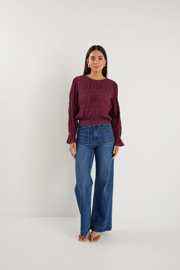 Woman wearing a long-sleeve textured blouse in deep plum, styled with blue high-waisted jeans and gold hoop earrings.