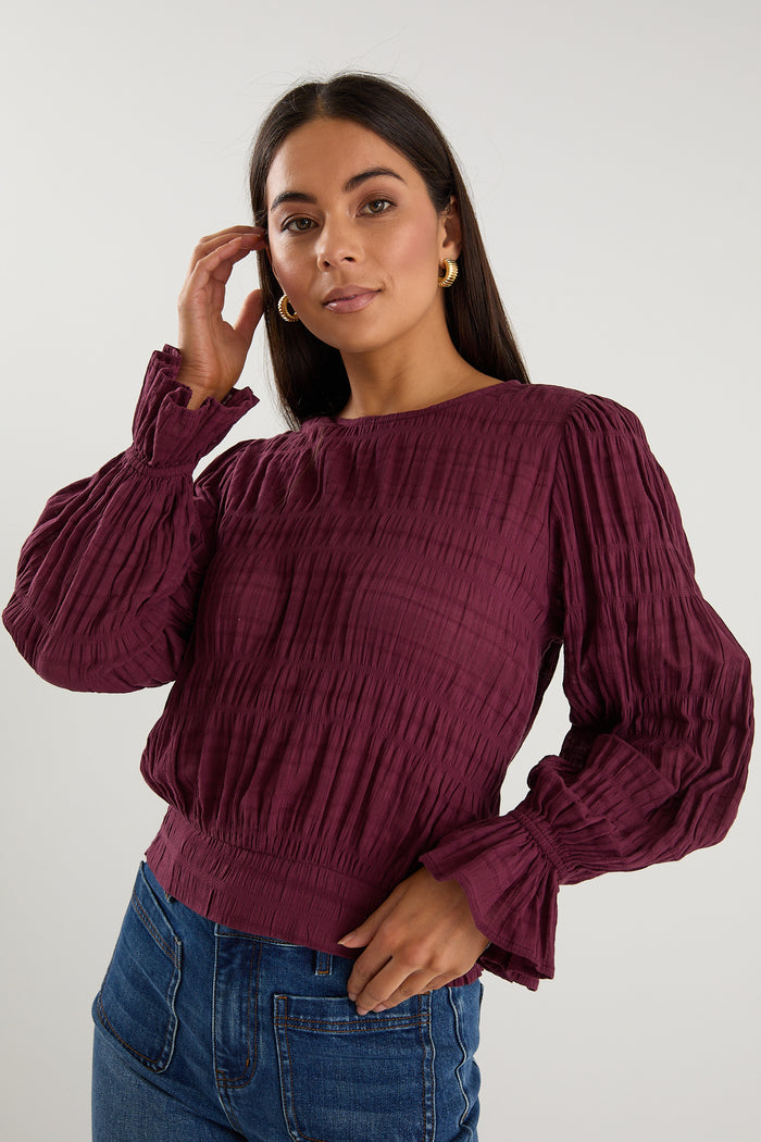 Woman wearing a long-sleeve textured blouse in deep plum, styled with blue high-waisted jeans and gold hoop earrings.