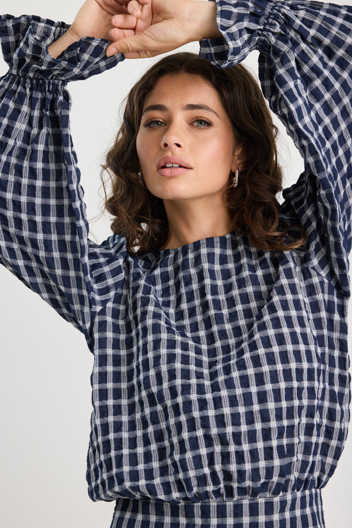 Woman wearing a blue and white checkered shirt with arms raised against a light background