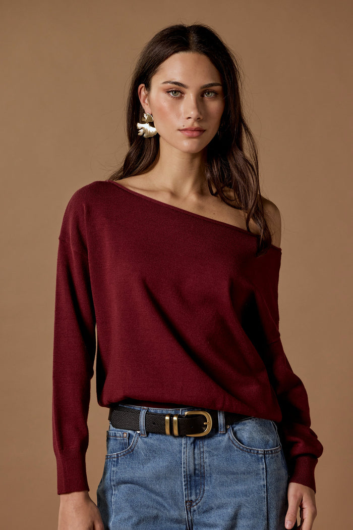 Woman wearing a burgundy off-shoulder top and blue jeans against a brown background