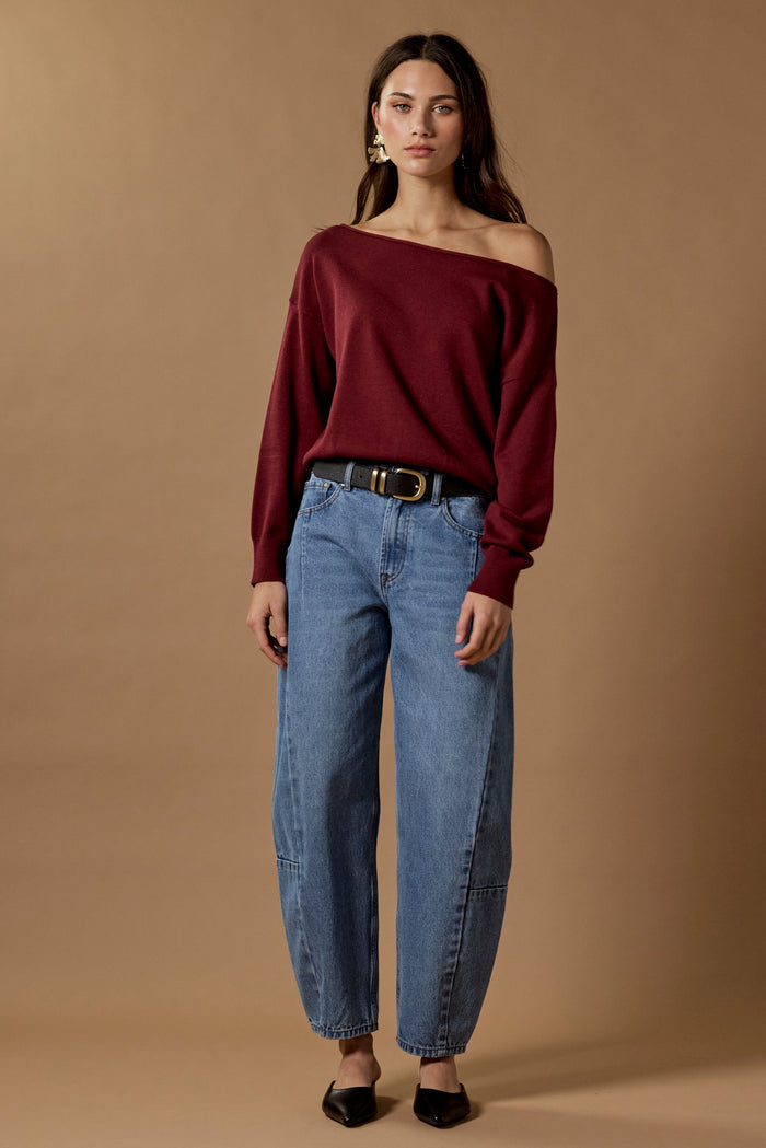 Woman wearing a burgundy off-shoulder top and blue jeans against a brown background