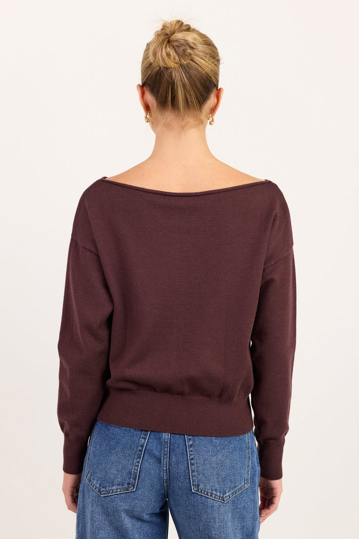A woman viewed from the back wearing a dark plum-brown wide-neck sweater and blue jeans against a plain off-white background.