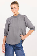 A woman modeling a gray and white micro-checkered blouse with a textured embroidered yoke and rolled three-quarter sleeves, paired with blue jeans against a plain white studio background.