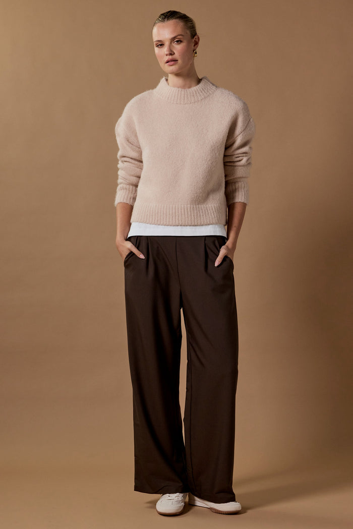 Person wearing a beige sweater and brown pants against a brown background
