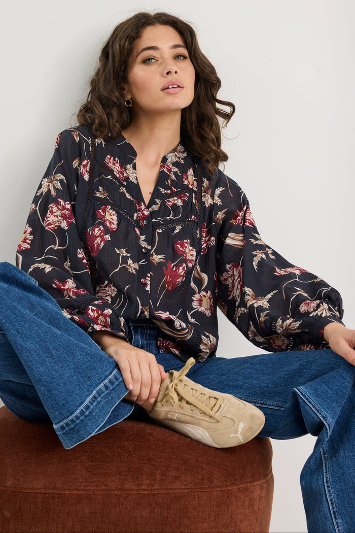 Woman wearing a floral blouse and blue jeans sitting on a brown ottoman against a white background
