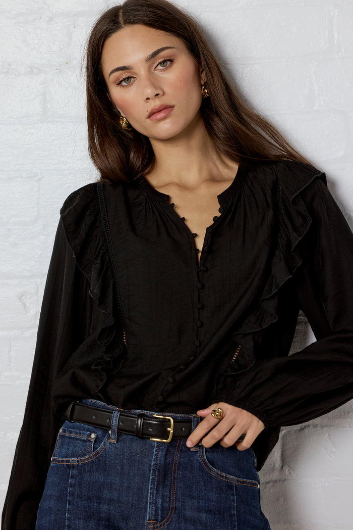 Woman wearing a black blouse with ruffled details and blue jeans against a white brick wall.