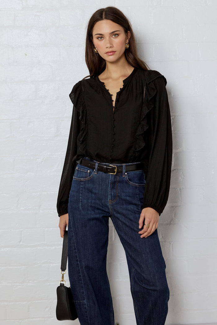 Woman wearing a black blouse and blue jeans against a white brick wall.