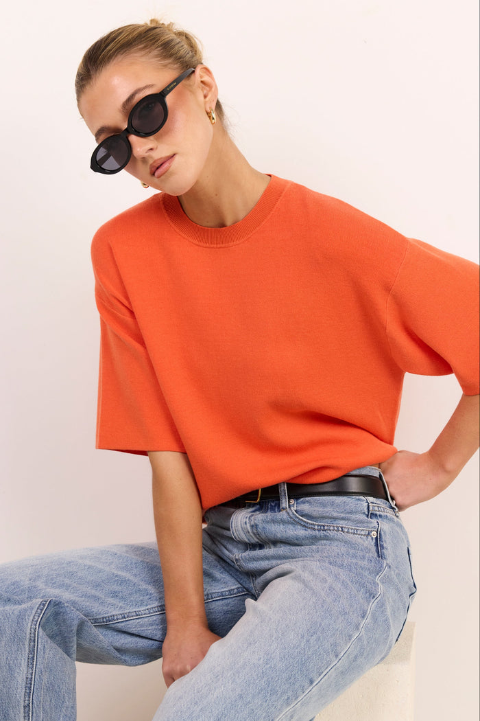 A woman wearing an oversized orange short-sleeve crewneck t-shirt and light-wash blue jeans, sitting against a plain off-white background.