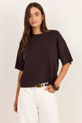 A woman modeling a dark brown short-sleeve knit top with white pants, standing against a plain off-white studio background.