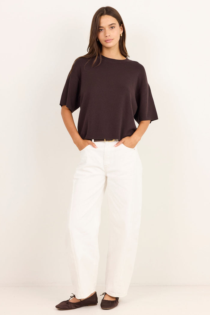 A woman modeling a dark brown short-sleeve top and white wide-leg pants against a plain white studio background.
