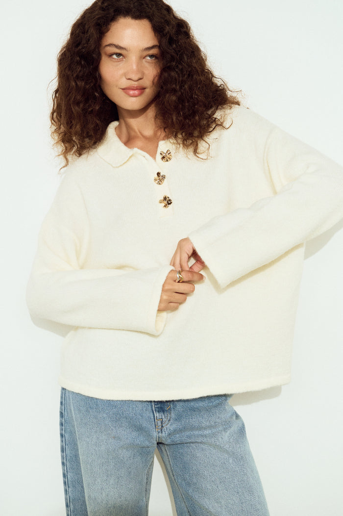 Woman wearing a cream sweater with gold buttons and blue jeans on a white background
