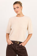 A woman modeling a short-sleeved, fuzzy cream-colored knit sweater paired with dark brown wide-leg jeans and a gold-buckled belt, set against a plain light gray studio background.