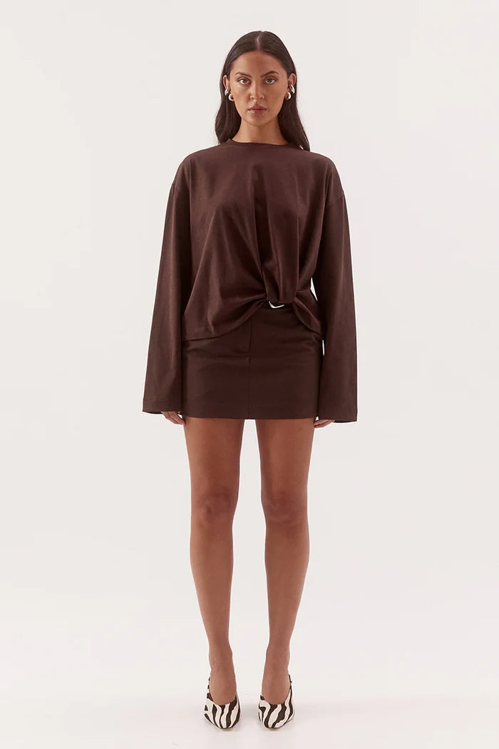 A model wearing a dark brown long-sleeve mini dress with a twisted knot detail at the waist, paired with black and white zebra-patterned heels, against a plain white studio background.