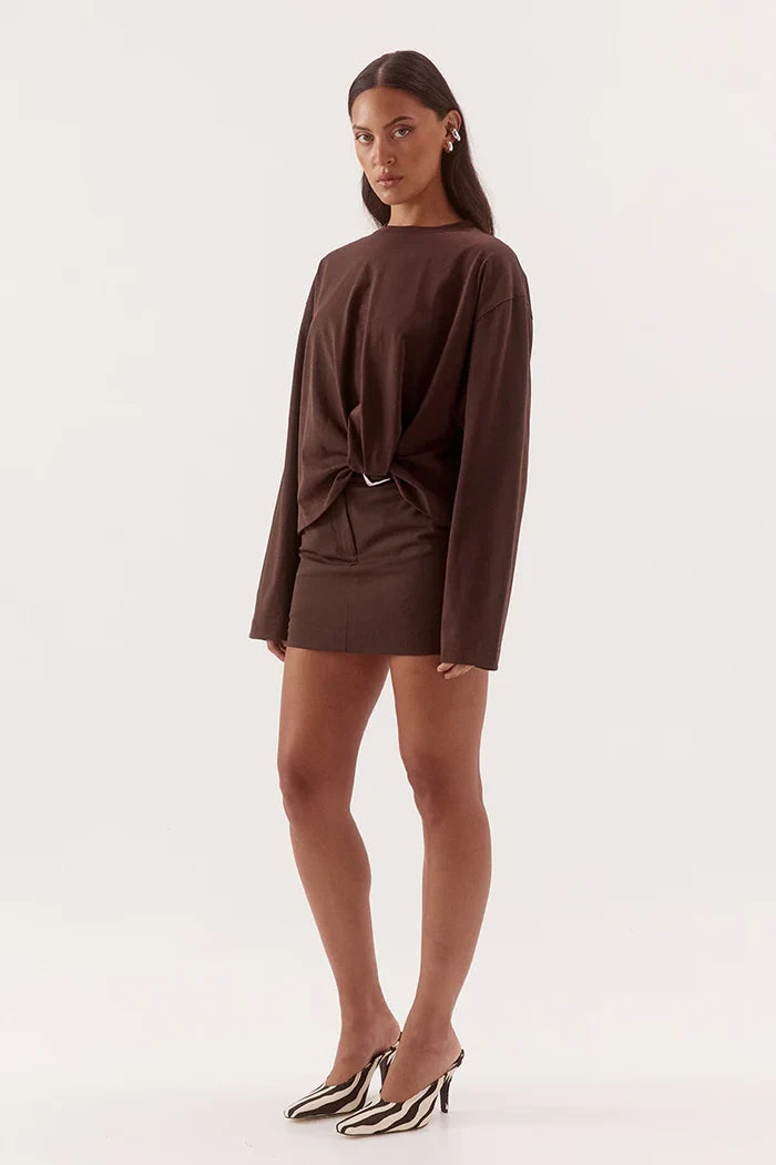 A model wearing a dark brown long-sleeve mini dress with a front knot detail and a thin belt, paired with black and white striped mule heels, against a plain white studio background.