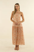 A woman modeling a sleeveless, tiered maxi dress with a small orange and green floral print on a light peach-beige base, set against a plain cream studio background.