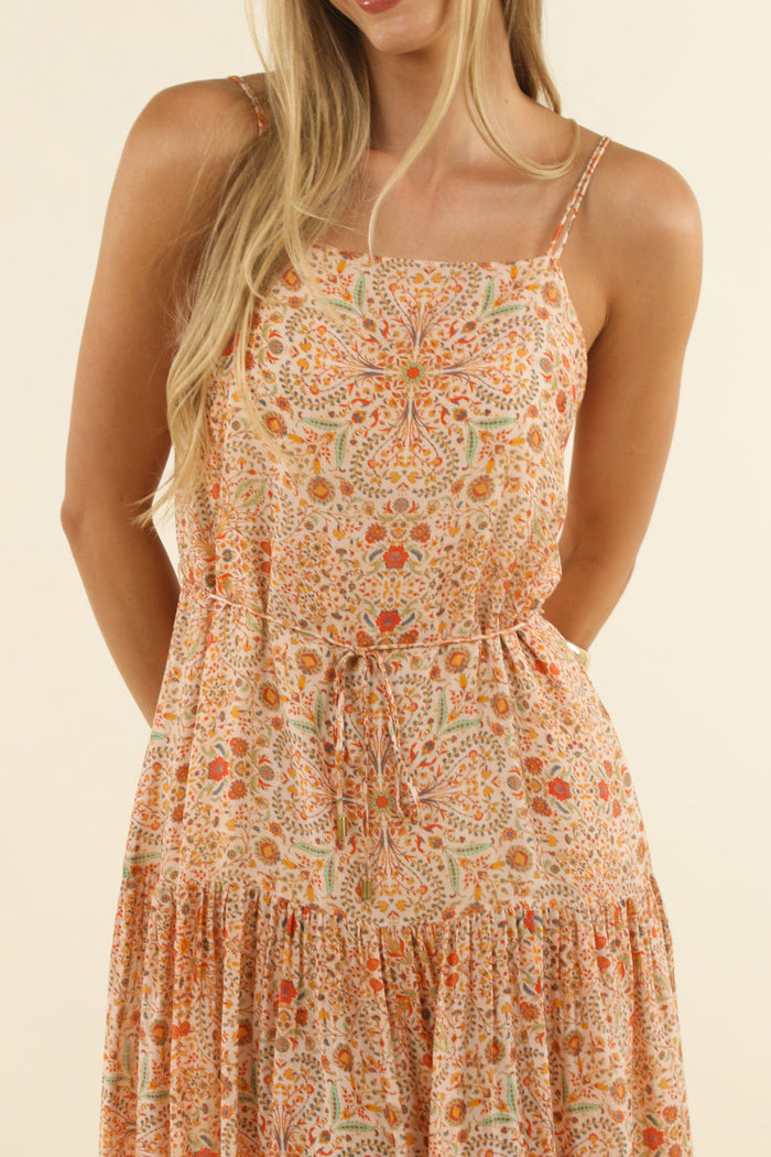 A woman wearing a sleeveless tiered dress with a dense orange, peach, and beige floral mandala pattern against a solid cream background.