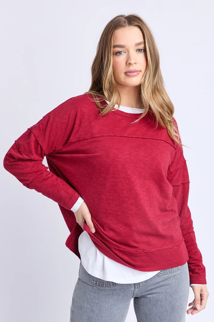 Woman wearing a red sweater over a white shirt with gray pants on a light gray background