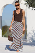 A woman walking outdoors near a white architectural structure and pool, wearing a brown sleeveless top and a long brown-and-beige checkered skirt, with a brown handbag.