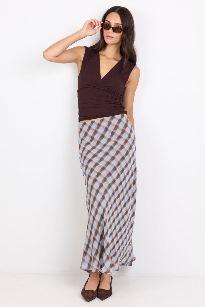 A woman modeling a sleeveless dark brown wrap-style top and a long brown, white, and light blue gingham skirt, paired with brown heels and sunglasses, against a plain white studio background.