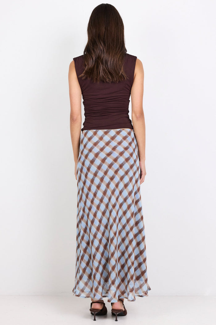 A woman is shown from behind wearing a sleeveless dark brown top and a long skirt with a brown, beige, and light blue gingham pattern, standing against a plain white studio background.
