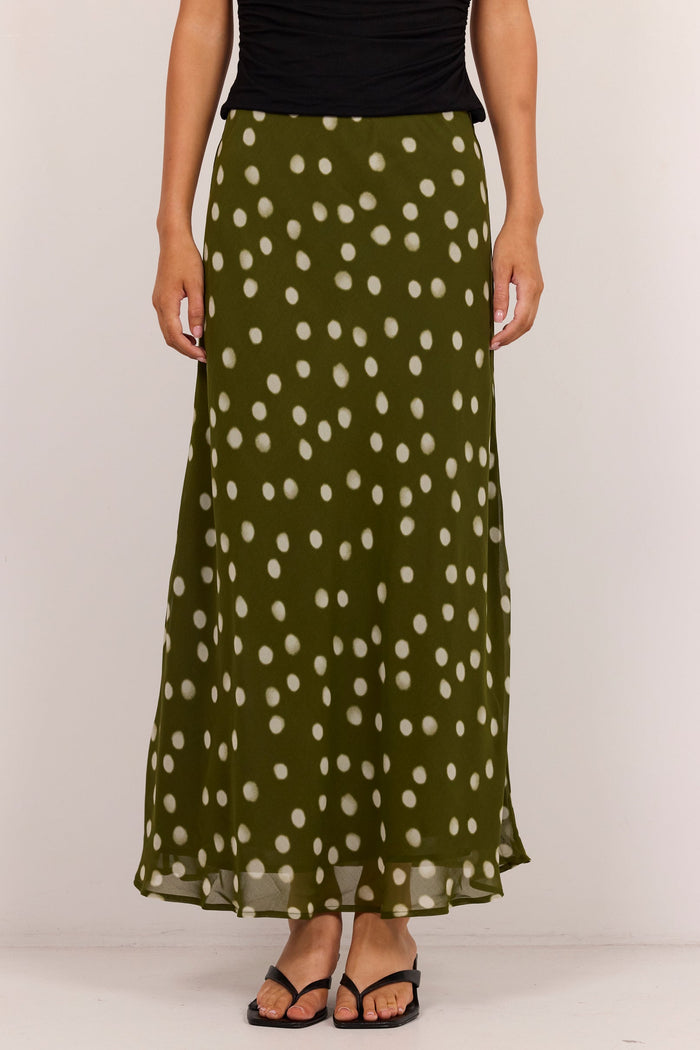 An olive green maxi skirt with a white polka dot pattern, worn by a model with a black top and black sandals against a plain white studio background.