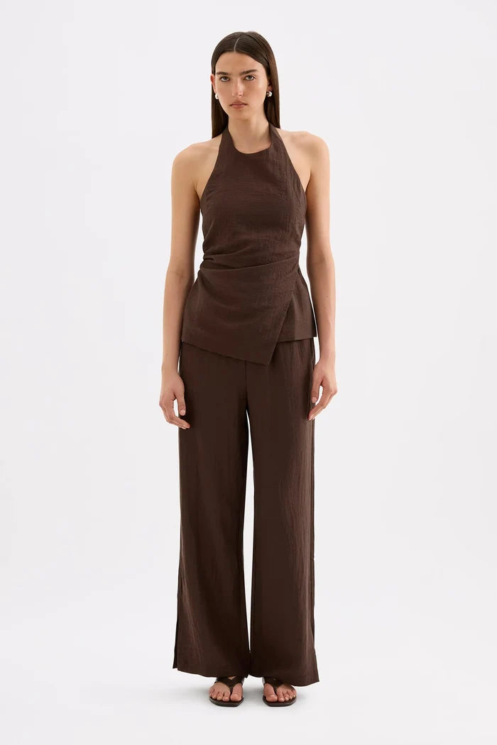 A model stands against a light gray studio background wearing a dark brown sleeveless halter top with a draped wrap-style waist and matching wide-leg pants.