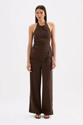 A model stands against a light gray studio background wearing a dark brown sleeveless halter top with a draped wrap-style waist and matching wide-leg pants.