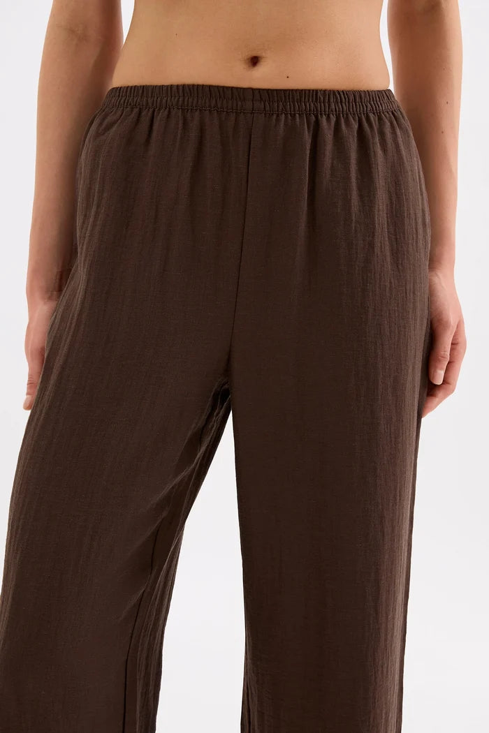 A close-up of a person wearing loose, wide-leg brown pants with an elastic waistband, shown from the torso down against a plain light background.
