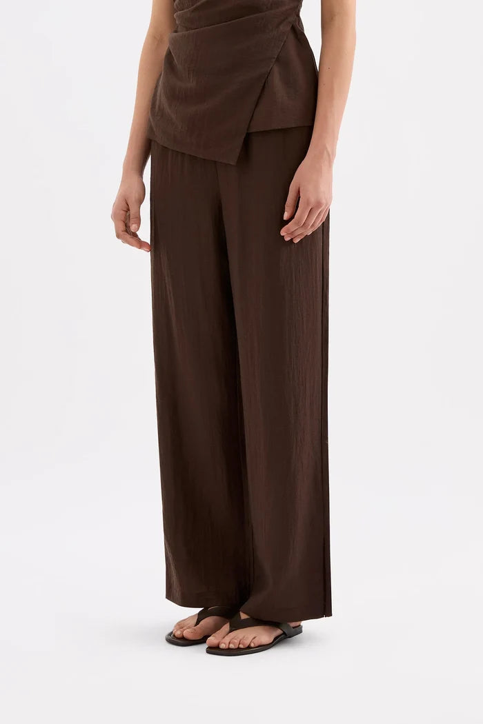 A model wearing dark brown wide-leg trousers and a matching dark brown wrap-style top, paired with black flat sandals, against a plain white studio background.