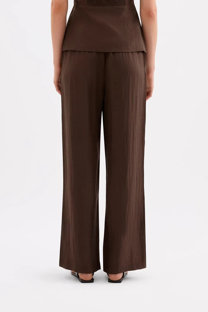 A model shown from the back wearing dark brown wide-leg pleated trousers and a matching dark brown top against a plain white studio background.