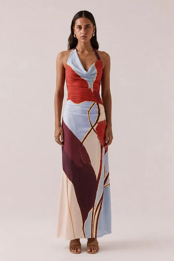 A woman modeling a halter-neck maxi dress with an abstract geometric print in red, light blue, cream, and burgundy with gold lines, set against a solid light beige background.