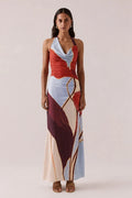 A woman modeling a halter-neck maxi dress with an abstract geometric print in red, light blue, cream, and burgundy with gold lines, set against a solid light beige background.