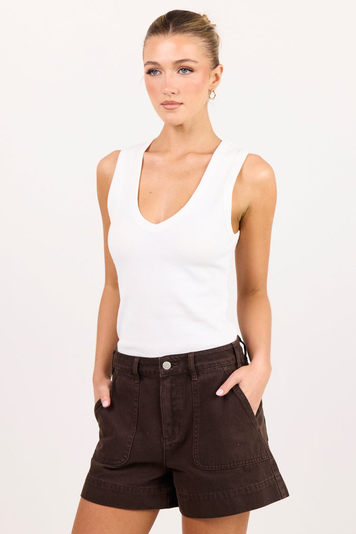 A woman modeling a white ribbed sleeveless V-neck tank top and dark brown high-waisted shorts against a plain white studio background.