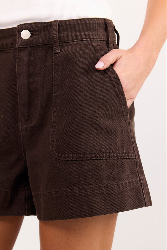 Close-up of a person wearing dark brown high-waisted shorts with front patch pockets and a white top, against a plain light background.