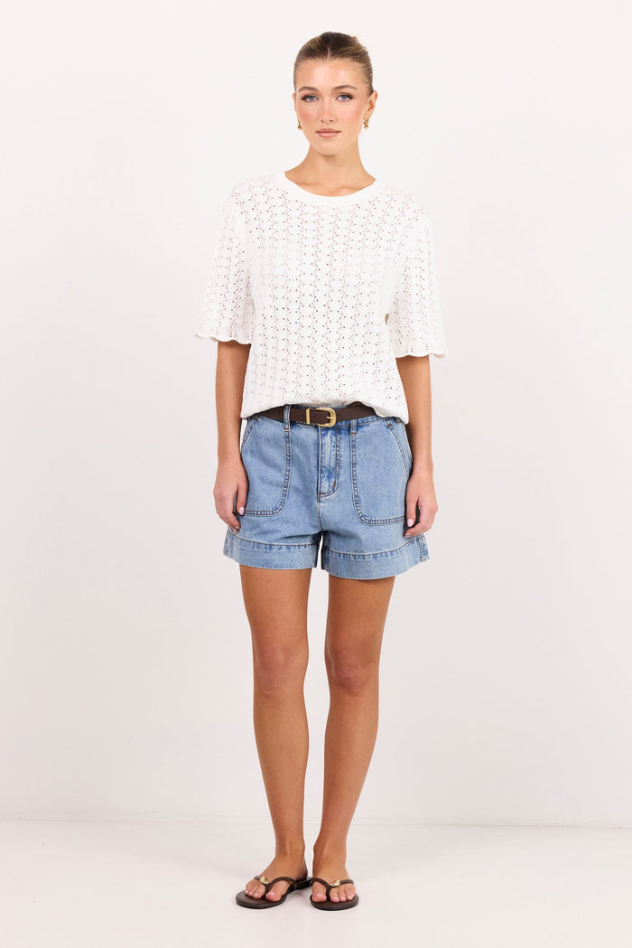 A woman modeling a white textured short-sleeve top and light blue denim shorts with a brown belt, standing against a plain white studio background.