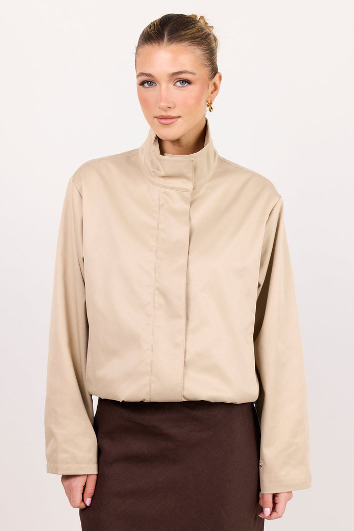 A woman modeling a beige long-sleeve mock-neck jacket with a unique front seam detail, paired with a dark brown skirt, against a plain white studio background.