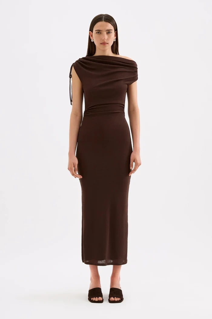 A woman modeling a dark brown off-the-shoulder midi dress with a draped neckline and side tie detail, paired with dark brown heeled sandals, against a plain white studio background.