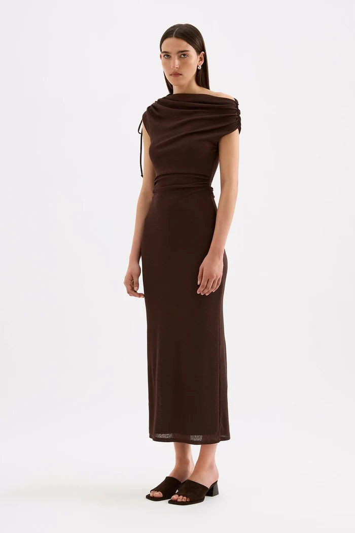 A woman modeling a dark brown off-the-shoulder midi dress with a draped neckline, side tie detail, and a fitted silhouette, paired with dark brown slide sandals against a plain white studio background.