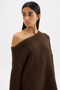 A woman modeling a dark brown, chunky knit off-the-shoulder sweater against a plain light gray studio background.