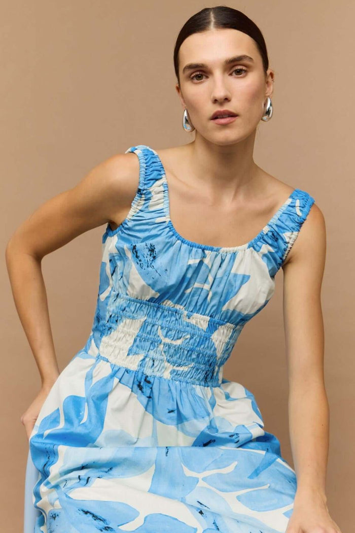 Woman wearing a light blue and white sleeveless midi dress with a shirred waist and soft leaf print, featuring a relaxed, floaty silhouette.