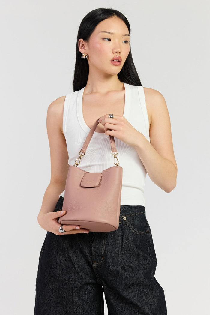 A model wearing a white tank top and dark wide-leg jeans holds a dusty pink leather bucket bag with gold-tone hardware against a light gray studio background.