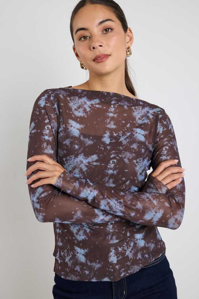 Woman wearing a long sleeve brown and blue tie-dye mesh top with dark jeans, standing with hands in pockets