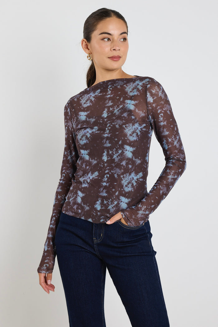 Woman wearing a long sleeve brown and blue tie-dye mesh top with dark jeans, standing with hands in pockets