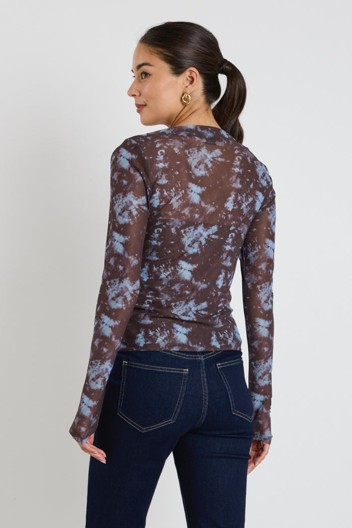 Woman wearing a long sleeve brown and blue tie-dye mesh top with dark jeans, standing with hands in pockets