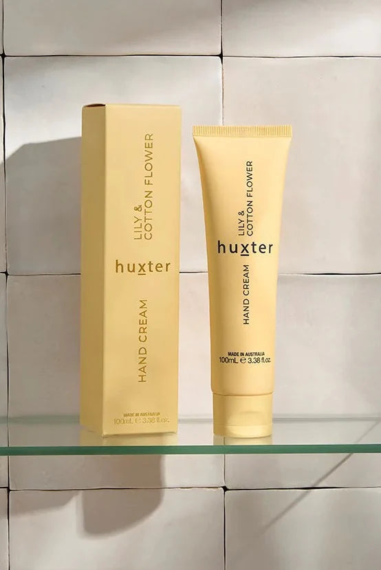 A yellow tube of Huxter hand cream in the scent "Lily & Cotton Flower" stands beside its matching rectangular box on a clear glass shelf, against a soft-tiled beige wall with natural light casting gentle shadows.